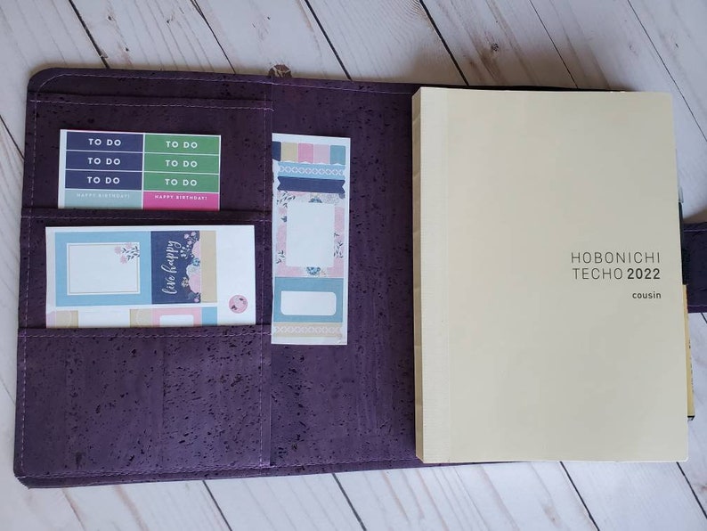 May include: Open purple cork planner with a cream-colored Hobonichi Techo 2022 cousin notebook. The planner has pockets holding colorful sticky notes with "To Do" and "Happy Birthday" labels, and floral designs. The planner is open.