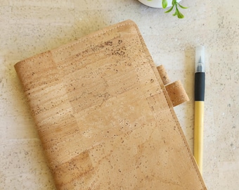 Op de afbeelding: Een lichtbruin notitieboek van kurk met een bijpassende tabsluiting. Een zwarte en bruine pen ligt naast het notitieboek. Een kleine potplant met groene bladeren staat op de achtergrond. Het notitieboek is gemaakt van natuurlijk materiaal.