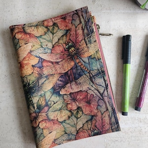 May include: A zippered wallet with a dragonfly and leaf pattern in shades of pink, orange, blue, and green. Two pens, one green and one purple, are next to the wallet. The wallet has a pink zipper.