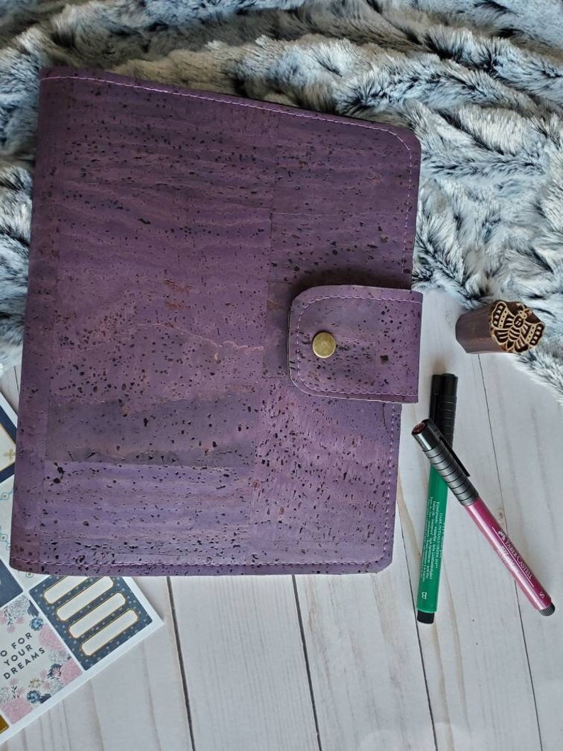 May include: A purple cork journal with a snap closure and a gold button. The journal is next to a wooden stamp, two pens, and a small sticker sheet with the text "GO FOR YOUR DREAMS". The journal is on a white wooden surface.