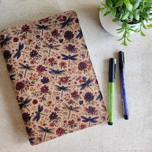 Op de afbeelding: Een notitieboek met een bloemen- en libellenpatroon in tinten bordeauxrood, paars en beige. Twee pennen, een groene en een paarse, liggen naast het notitieboek. Een kleine potplant staat op de achtergrond.