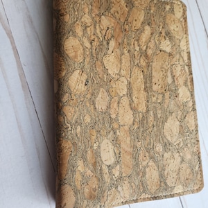 May include: A rectangular cork notebook cover with a natural, textured pattern. The cover is stitched with white thread along the edges. The notebook is closed and appears to be new.