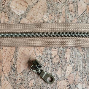 May include: Close-up of a tan zipper with a silver metal zipper pull on a cork surface. The zipper teeth are silver, and the fabric is a solid tan color. The zipper pull is a bronze color.