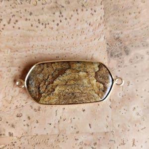 Puede incluir: Un colgante de forma ovalada con una piedra de jaspe paisaje pulida en tonos marrón y beige, engastada en un fino marco de metal dorado. El colgante incluye un pequeño aro metálico circular en cada uno de los dos extremos opuestos.