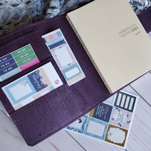 May include: A purple cork planner with a cream-colored Hobonichi Techo 2022 Cousin notebook. The planner contains sticker sheets and sticky notes with floral and geometric designs. The stickers include the phrases "To Do" and "Happy Birthday!"