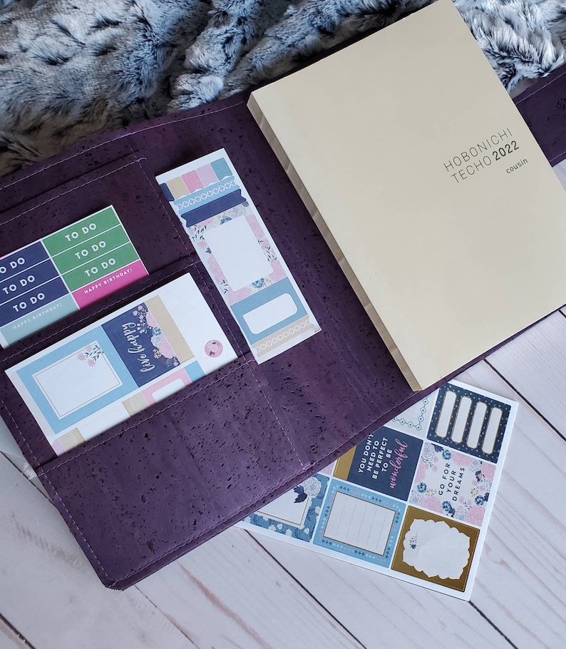 May include: A purple cork planner with a cream-colored Hobonichi Techo 2022 Cousin notebook. The planner contains sticker sheets and sticky notes with floral and geometric designs. The stickers include the phrases "To Do" and "Happy Birthday!"