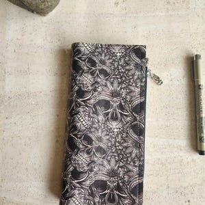 May include: A rectangular wallet featuring a black and white intricate pattern of skulls and floral motifs. The wallet has a side zipper closure with a silver-toned metal pull. A black Micron pen is positioned to the right of the wallet.