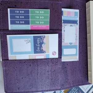May include: A purple cork planner with pockets holding colorful stickers and a bookmark. The stickers include "TO DO" labels in blue and green, and "HAPPY BIRTHDAY!" labels in pink. The bookmark has pastel designs. Additional stickers with text are visible.