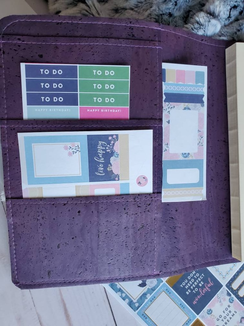 May include: A purple cork planner with pockets holding colorful stickers and a bookmark. The stickers include "TO DO" labels in blue and green, and "HAPPY BIRTHDAY!" labels in pink. The bookmark has pastel designs. Additional stickers with text are visible.