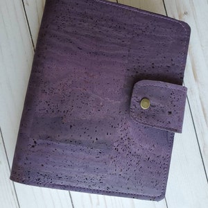 May include: A purple cork journal with a snap closure. The journal has a textured surface and a stitched border. The snap closure is secured with a gold-colored button. The journal is closed and ready to be used.