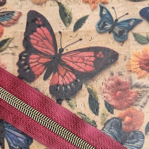 May include: A cork fabric sheet featuring a printed pattern of colorful butterflies and flowers in shades of red, blue, and orange, overlaid with a diagonal strip of deep red fabric featuring a metallic gold-toned zipper track.