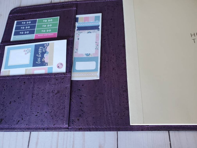 May include: A purple cork planner with multiple pockets holding stickers and a notepad. The stickers have text that says "TO DO" and "Happy Birthday!" The notepad is beige. The planner is open, revealing the interior.