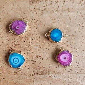 May include: Four gemstone pendants with gold-colored metal edges and loops. The gemstones are in shades of blue and purple, with a natural, textured appearance. The pendants are displayed on a textured, light brown surface.