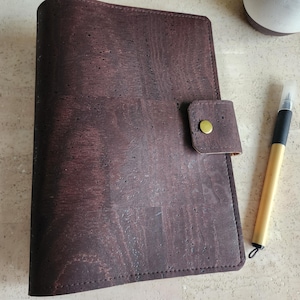 May include: A dark brown cork journal with a flap closure and a gold button. A pen with a black and gold barrel rests beside the journal. The journal's cover has a textured appearance.