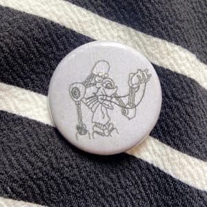 Può includere: Un bottone rotondo e grigio con uno schizzo di una figura simile a un robot. La figura ha una testa scheletrica e braccia e gambe meccaniche. Il bottone è posto su uno sfondo a righe bianche e nere.