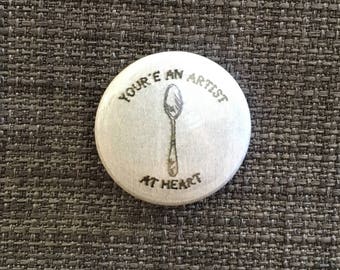 Könnte beinhalten: Ein weißer runder Knopf mit dem Text "You're an artist at heart" und einer Zeichnung eines Löffels in der Mitte.
