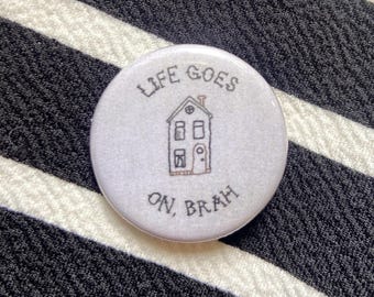 Könnte beinhalten: Ein runder, weißer Button mit dem Text "LIFE GOES ON, BRAH" in schwarzer Schrift. Eine kleine Hausillustration ist auf dem Button zentriert. Der Button befindet sich vor einem schwarz-weiß gestreiften Hintergrund.