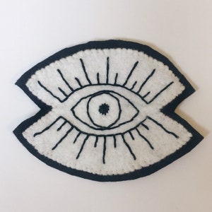 May include: A white felt eye patch with a black embroidered eye and eyelashes. The eye patch is surrounded by a black felt border.