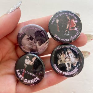 May include: Four circular pin-back buttons featuring photographic prints of musicians. The buttons display text including "Why can't we give love that one more chance?", "Can't we give ourselves one more chance?", "This is our LAST DANCE", and "This is ourselves UNDER PRESSURE".