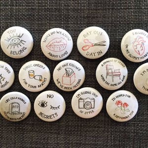 May include: A collection of 12 round pins with various illustrations and text. The pins feature a variety of designs, including a heart, an eye, a pair of scissors, a tombstone, a glass of wine, a tissue box, a chair, and a camera. The text on the pins includes phrases such as "You Belong", "What Do We Know About Love?", "Day Out Day In", "Waiting For This To Make Sense", "Three Cheers For Your Apathy", "No One Knows We've Been Crying", "A Change Of Heart With The Same Old Ache", "I Always Trip On My Clumsy Heart", "I'm Better Than No One", "Life Kills Rebels Death Gets The Rest", "No Secrets", "I'm Going Out Of Style", and "I'd Hoped For A Different You".