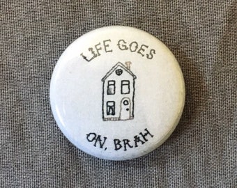 Könnte beinhalten: Weißer Knopf mit schwarzem Text, der "Life Goes On, Brah" lautet, und einer Schwarzweißillustration eines Hauses.