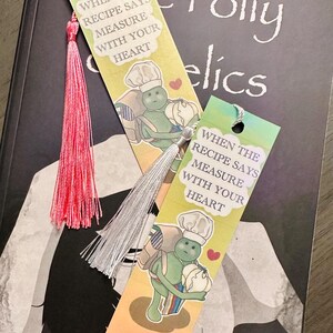 May include: Two colorful bookmarks with tassels rest on a book. Each bookmark features a cartoon chef holding a vegetable and the text "WHEN THE RECIPE SAYS MEASURE WITH YOUR HEART." One tassel is pink, the other is silver.