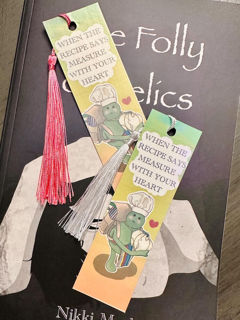 May include: Two colorful bookmarks with tassels rest on a book. Each bookmark features a cartoon chef holding a vegetable and the text "WHEN THE RECIPE SAYS MEASURE WITH YOUR HEART." One tassel is pink, the other is silver.