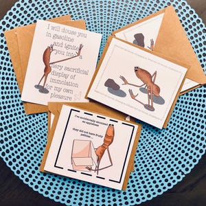 May include: Three greeting cards with brown envelopes. Each card features a cartoon character with humorous text. One card reads "I will douse you in gasoline and ignite you into a fiery sacrificial display of immolation for my own pleasure."