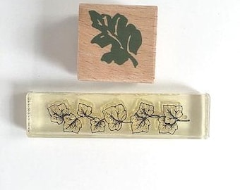 May include: Two wooden stamps with leaf designs. One stamp features a single green leaf, while the other stamp features a row of black leaves on a light yellow background.