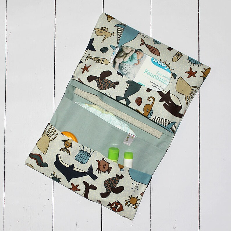 May include: A light blue and beige changing mat with a sea creature design. The mat is folded open to reveal a nappy, wipes, and two small bottles. The text "Sensitiv Feuchtt&uuml;cher" is visible on the wipes package.