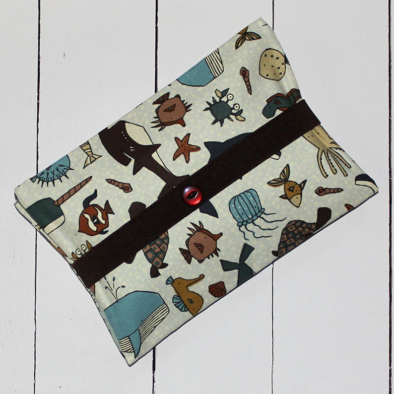 May include: A white fabric pouch with a brown strap and a red button closure. The pouch is decorated with a colourful sea life pattern, including fish, whales, jellyfish, and seahorses.