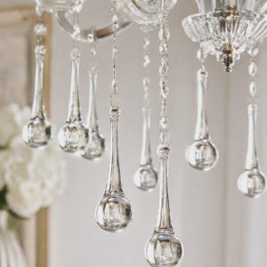 May include: A clear glass chandelier featuring multiple hanging teardrop-shaped crystals suspended from delicate metal chains. The crystals are faceted and transparent, catching light to create a reflective appearance throughout the fixture.