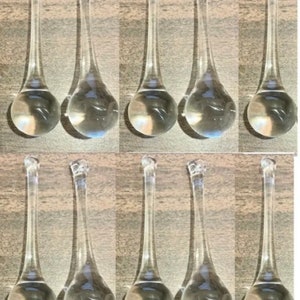 May include: Ten clear glass teardrop-shaped ornaments with a faceted top. The ornaments are transparent and reflect light, creating a sparkling effect. They are arranged in two rows of five on a neutral-toned surface.