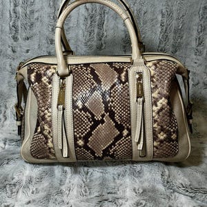 Peut inclure: Un sac à main structuré avec un panneau central à motif peau de serpent beige et marron, encadré par des sections latérales beiges unies. Le sac comprend deux poignées supérieures, une fermeture éclair et deux poches verticales zippées dorées.