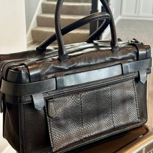 May include: A black leather handbag featuring a textured grain finish, two top handles, and a horizontal strap across the front. The bag includes a front pocket with a snakeskin-like pattern and a zipper closure with a leather pull tab.