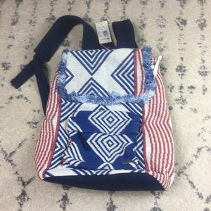 May include: A blue, red, and white patterned backpack with a frayed edge. The backpack has a large pocket on the front and adjustable straps.