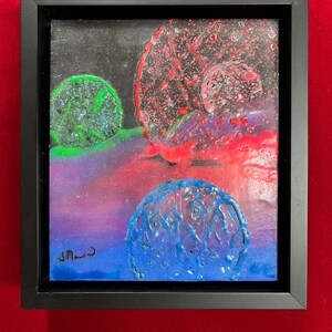 May include: A framed abstract painting with three circular shapes. The largest shape is red with black and white speckles. The middle shape is green with black speckles. The smallest shape is blue with white speckles. The background is a gradient of purple, pink, and red.