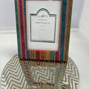 May include: A colorful striped picture frame made of recycled paper. The frame is handcrafted by Belmar Frame & Art and is for a 4x6 photo. The frame is sitting on a gold glitter chevron patterned surface.