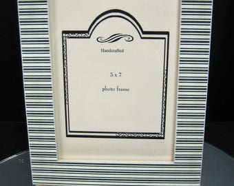 May include: A handcrafted 5x7 photo frame with a black and white striped border. The frame has a cream-colored interior with decorative black trim and the words "Handcrafted" and "5 x 7 photo frame" printed on the inside.