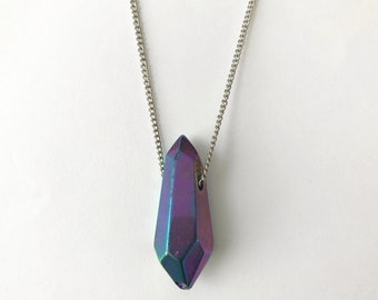 Op de afbeelding: Een zilveren ketting met een grote, regenboogkleurige, paarse kristallen hanger in de vorm van een traan.