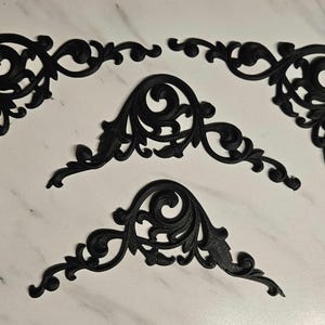May include: Set of black decorative corner pieces with ornate, swirling scrollwork designs. These architectural elements are likely used for embellishing furniture or interior decor, adding a touch of elegance and detail to any project.