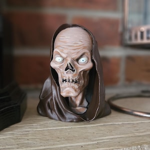 May include: A detailed, hand-painted skull sculpture with a brown hooded cloak. The skull has a pale complexion, a prominent nose, and glaring white eyes. The sculpture is a decorative item, likely for Halloween or gothic decor.