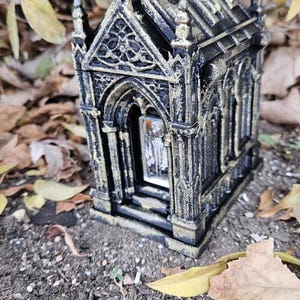 May include: A miniature black and gold gothic-style mausoleum with intricate architectural details. The small structure features arched doorways and windows, and is set against a backdrop of fallen leaves and foliage.