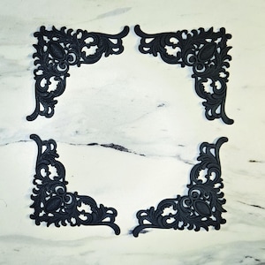 May include: Four black corner embellishments featuring intricate scrollwork and filigree patterns. Each piece has a symmetrical design with a central oval element and curved, leafy motifs extending outward to form a right-angle corner shape.