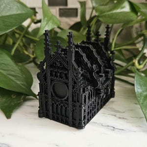 May include: A black, gothic-style miniature building with intricate architectural details. The model features pointed arches, spires, and decorative elements, creating a detailed and ornate appearance. The building is set against a backdrop of green foliage.