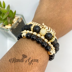 Peut inclure: Trois bracelets superposés avec des perles noires et dorées. Le bracelet du haut présente un charme en forme de feuille dorée avec un accent en cristal transparent. Le texte "Randi Gems" est visible sur le poignet.