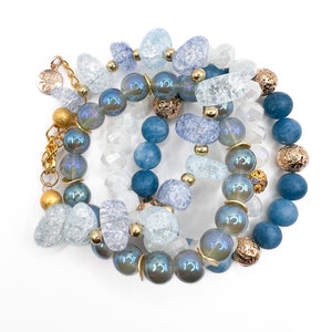May include: A set of four bracelets featuring blue and white glass beads, iridescent beads, and gold accents. The bracelets are layered and create a unique and stylish look.