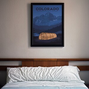 May include: Framed poster featuring a Colorado landscape with mountains, trees, and a glowing greenhouse. The poster has the word "COLORADO" at the top. The poster is hanging above a bed with a white pillow and blue sheet.