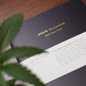 May include: A black grow planner with gold lettering that reads "Grow Planner Know Your Grow" and "Goldleaf Grow Planner". The planner has a white label with text that reads "Grow Better Cannabis. Whether you're a...", describing the planner's purpose.