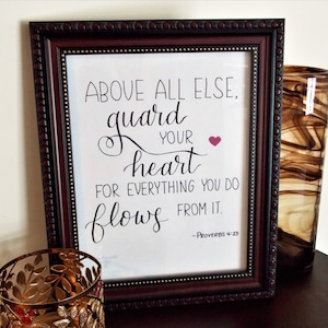 Może przedstawiać: Wydruk w ramie z czarno-białym cytatem kaligraficznym, który brzmi "ABOVE ALL ELSE, guard YOUR heart FOR EVERYTHING YOU DO flows FROM IT. -PROVERBS 4:23". Cytat jest napisany kursywą z małym czerwonym sercem nad słowem "heart".